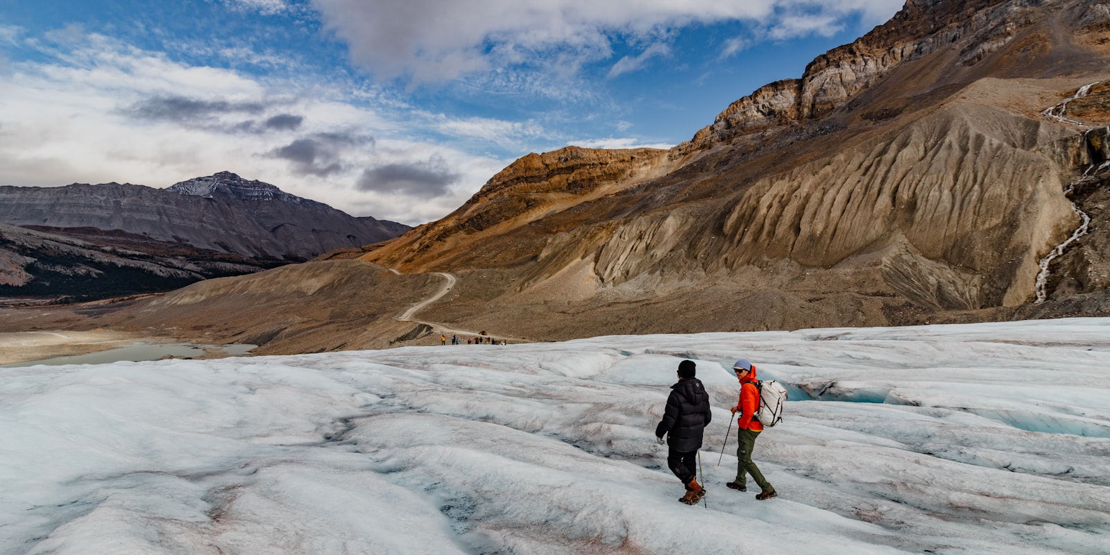 Two hikers trekking across a glacier toward snow-capped mountains